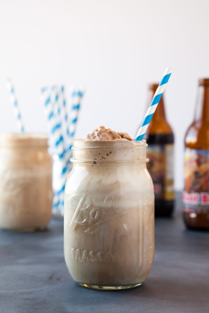 A root beer float with a striped straw, root beer bottles in the background.