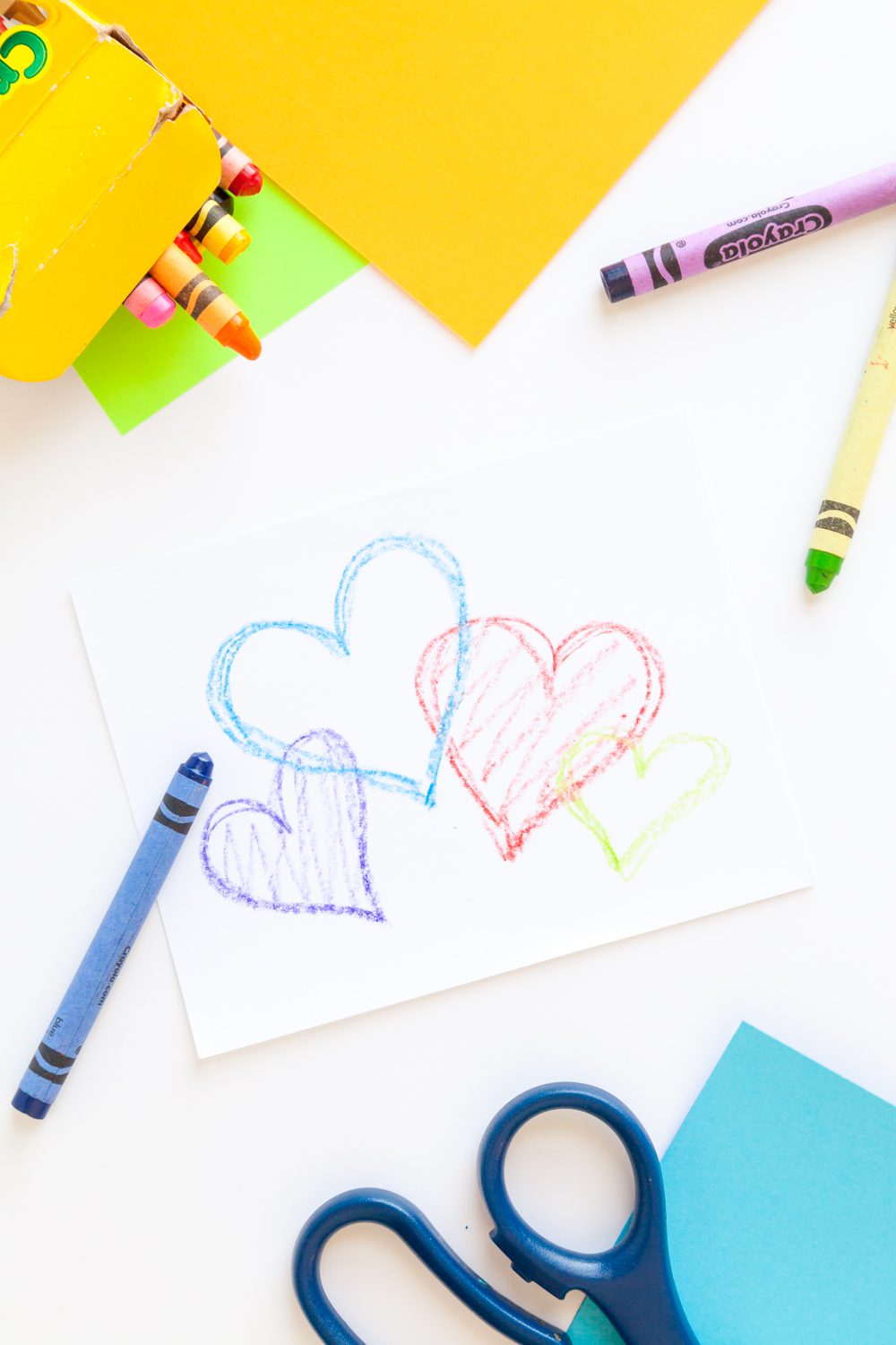 A card with colored hearts, surrounded by blue and yellow paper, and crayons.