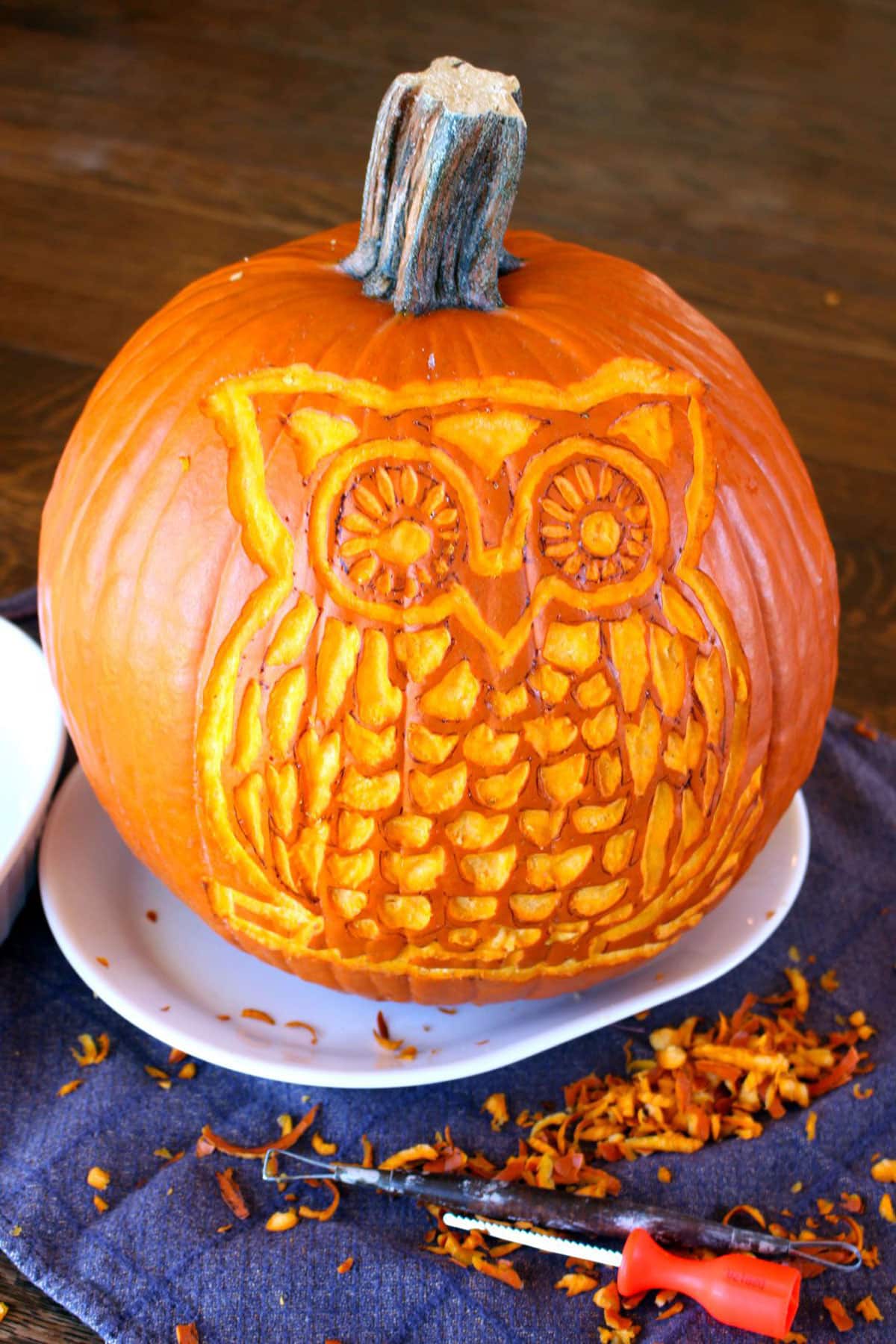 A woodland owl carved pumpkin sitting on a plate with carving tools and shavings, with a blue linen on a table.