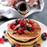 Pouring syrup over a stack of pancakes with nuts and berries on top.
