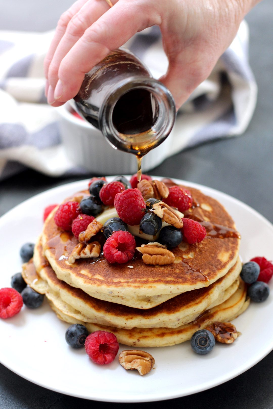 Pouring homemade syrup over a stack of pancakes with nuts, raspberries, and blueberries on top.