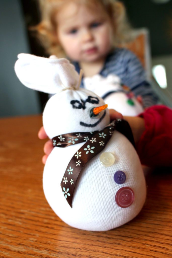 Child holding a sock snowman with ribbon, buttons, a nose, and face.