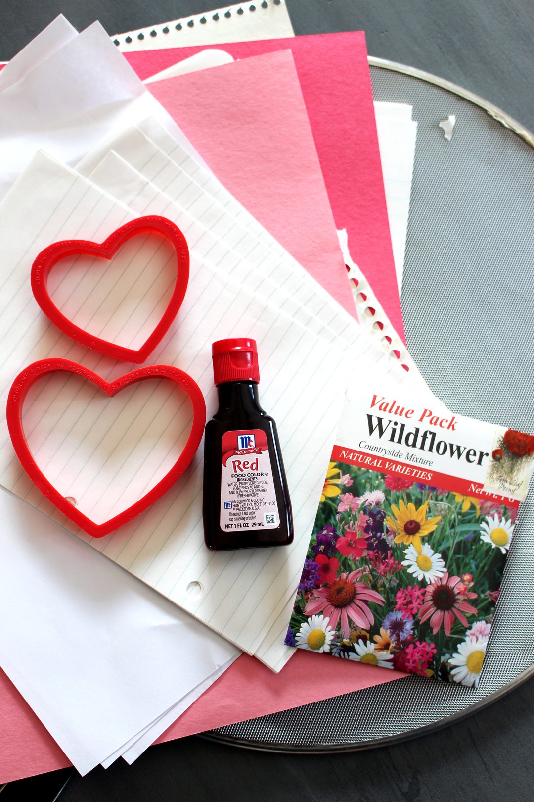 A pile of pink and white paper, red food coloring, wildflower seeds, heart cookie cutters, and a splatter screen.