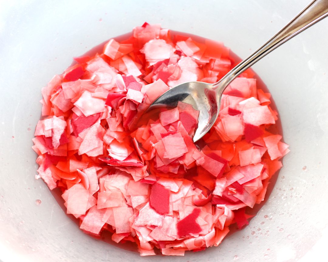 Paper scraps soaking in red water, stirred by a spoon.