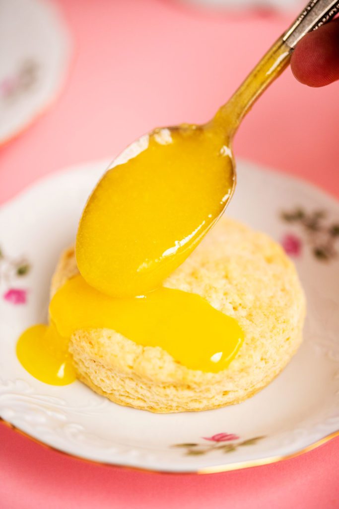 A spoon adding lemon curd to the top of a scone on a decorative plate.