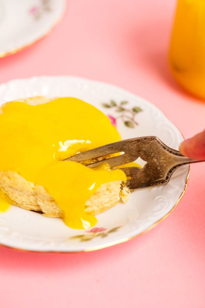 A fork taking a bite from a scone topped with lemon curd on a decorative plate.