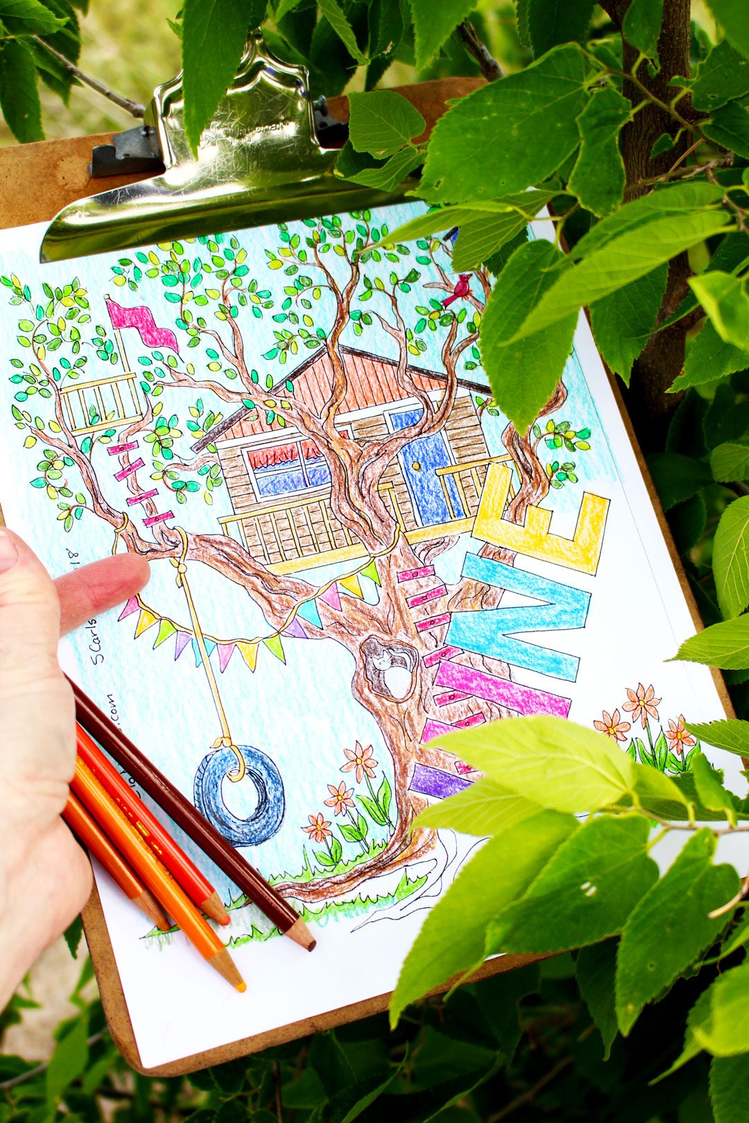 A hand holding colored pencils and a month of June coloring page with a tree house in a large tree.