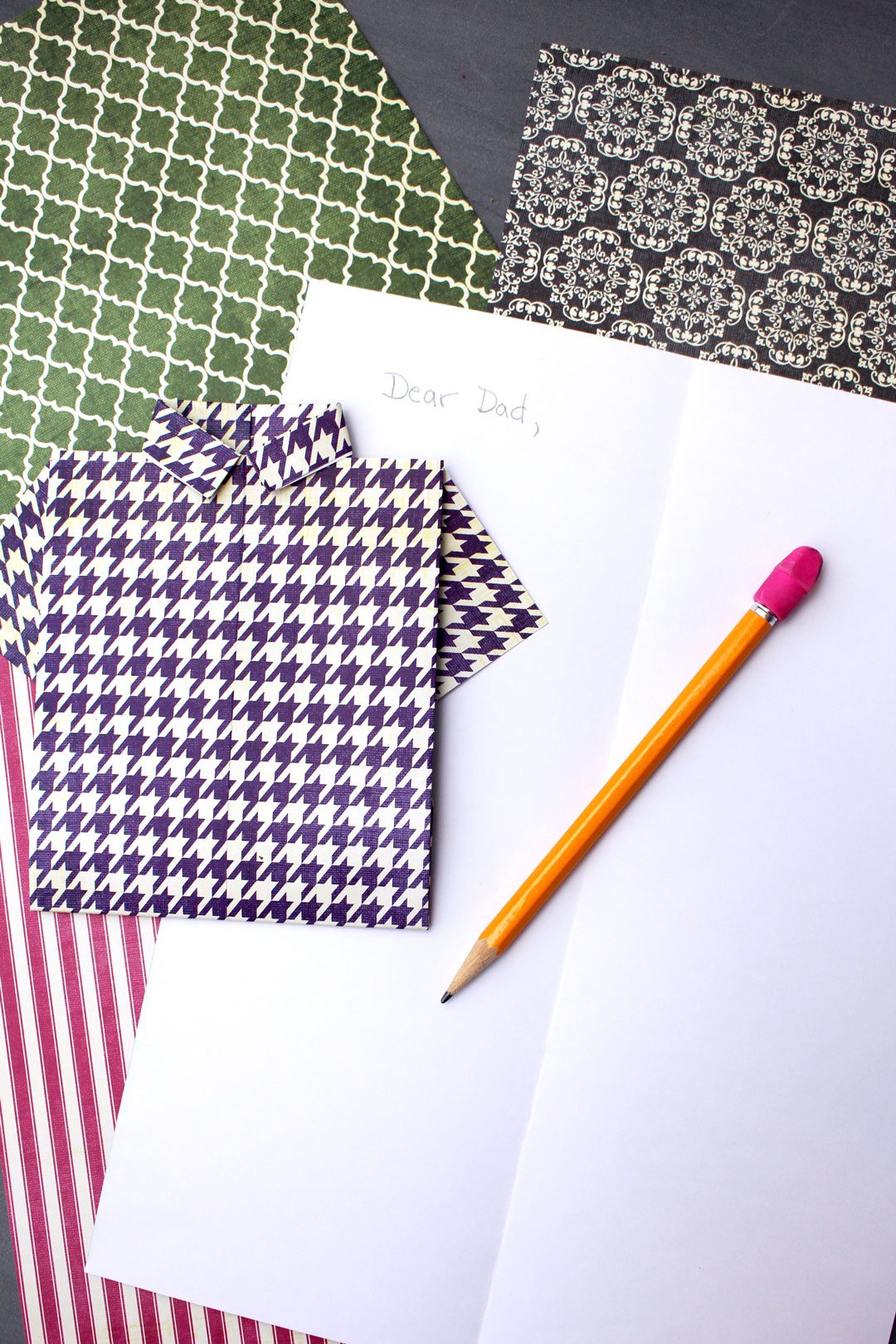 A folded origami shirt card next to several patterned scrapbook paper pieces, a pencil, and a note that reads, "Dear Dad,".