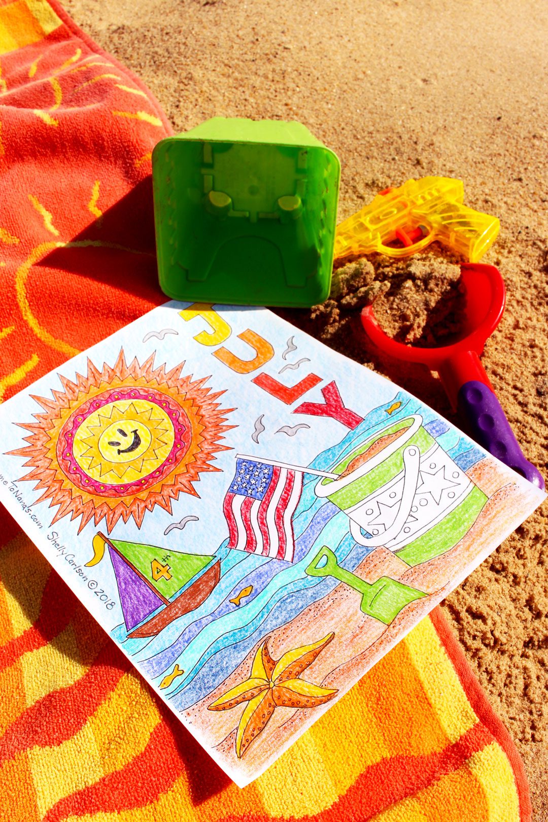 Finished July coloring page on beach towel on the sand with colored pencils near by.