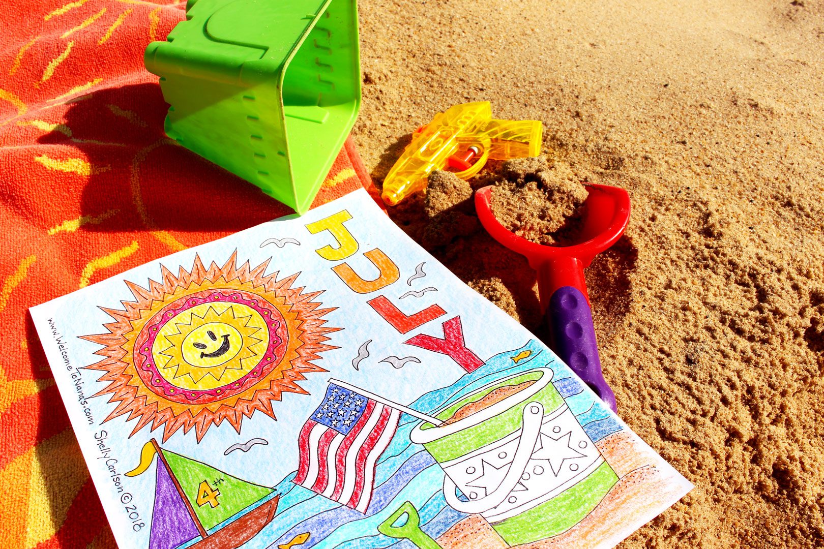 Finished July coloring page on beach towel on the sand with colored pencils near by.