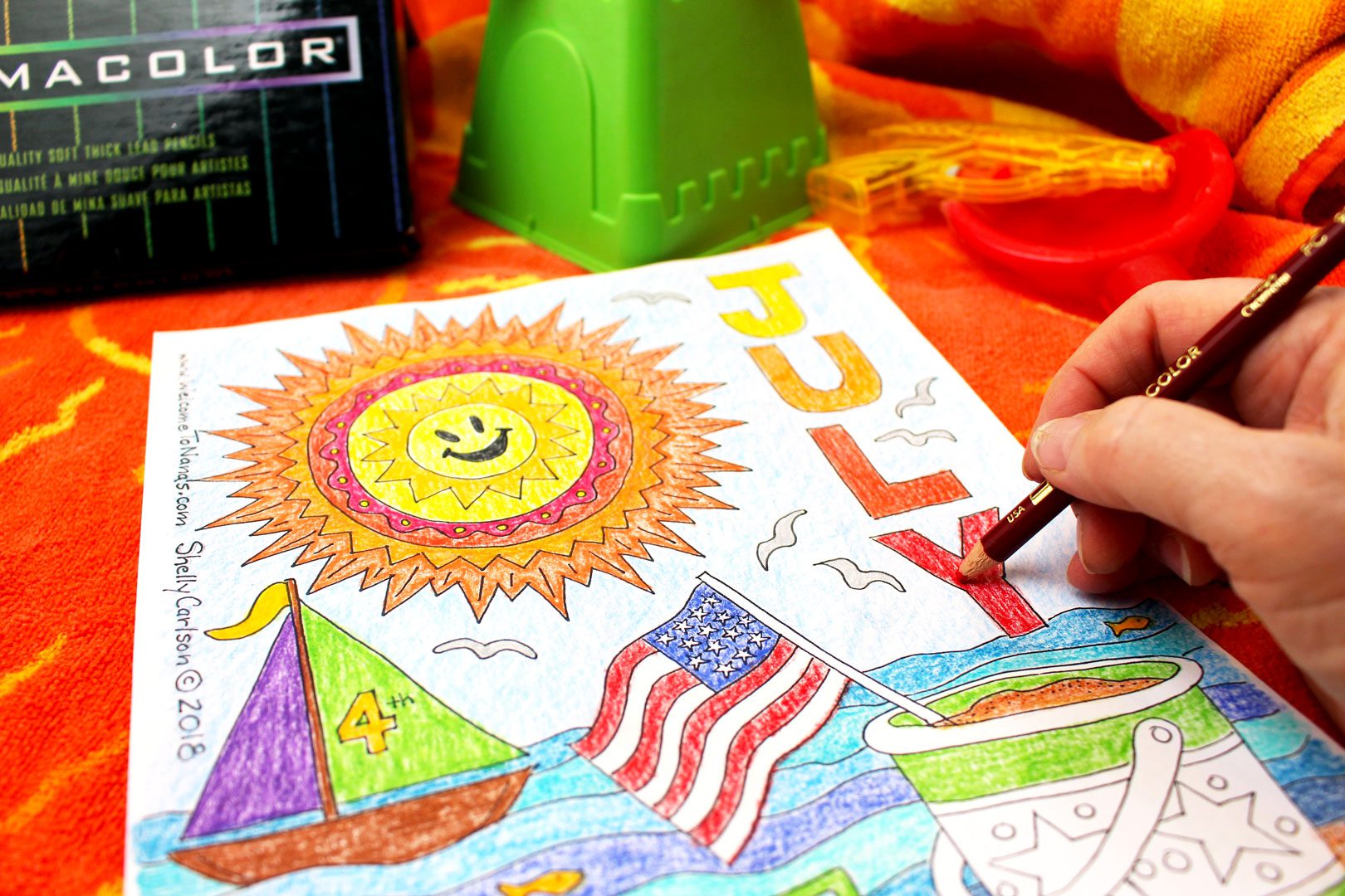 Coloring a July coloring page on beach towel on the sand with colored pencils near by.