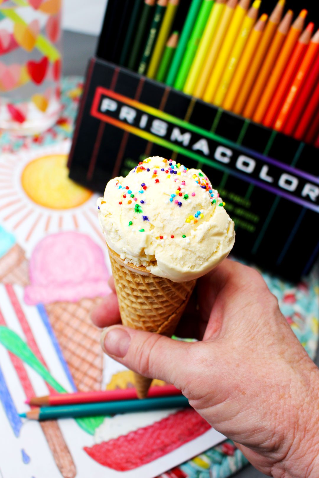 A hand holding an ice cream cone with a scoop of vanilla ice cream with sprinkles, in front of colored pencils and a coloring page.