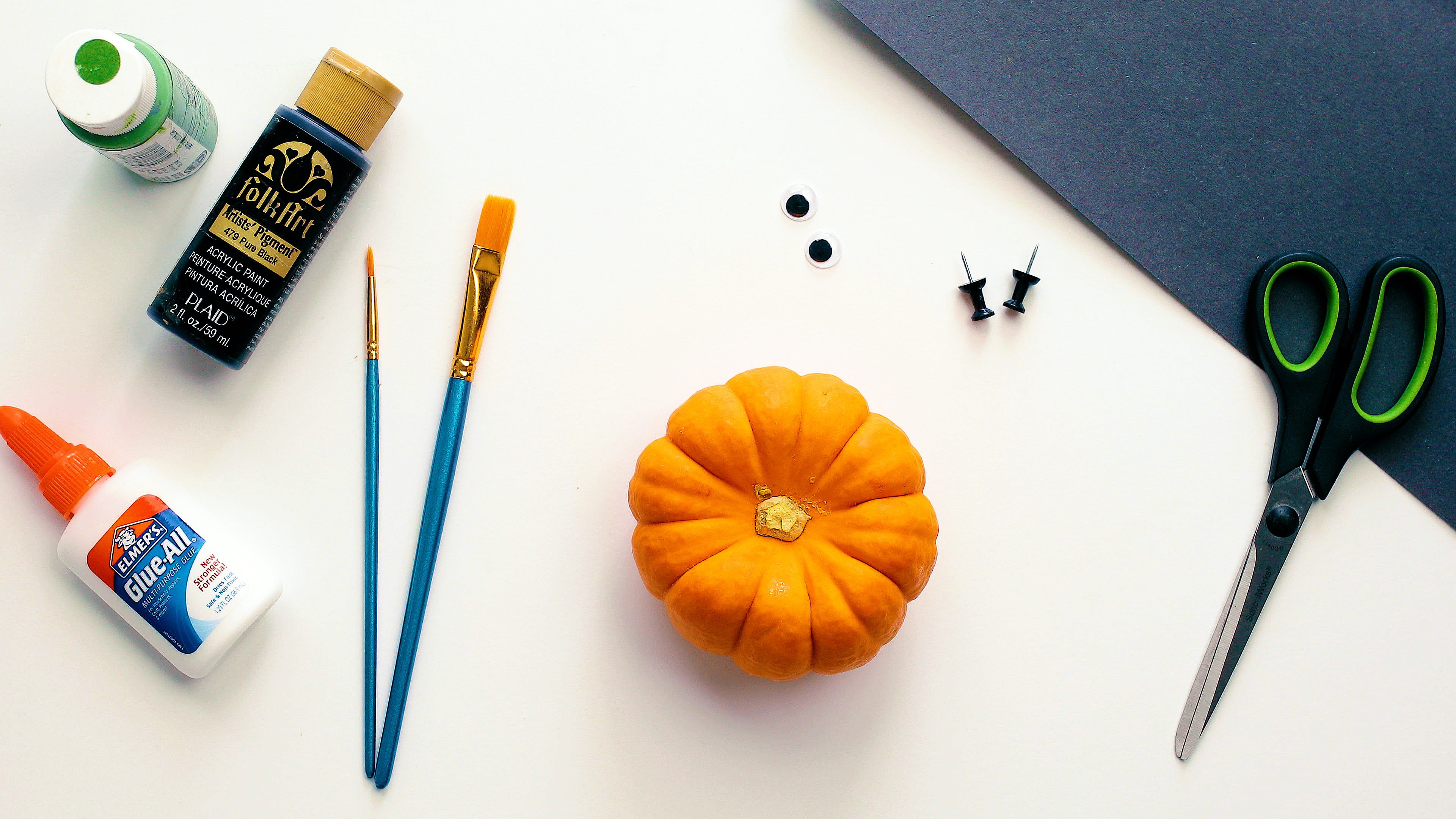 A mini pumpkin, paintbrushes, paint bottles, googly eyes, black construction paper, and push pins.