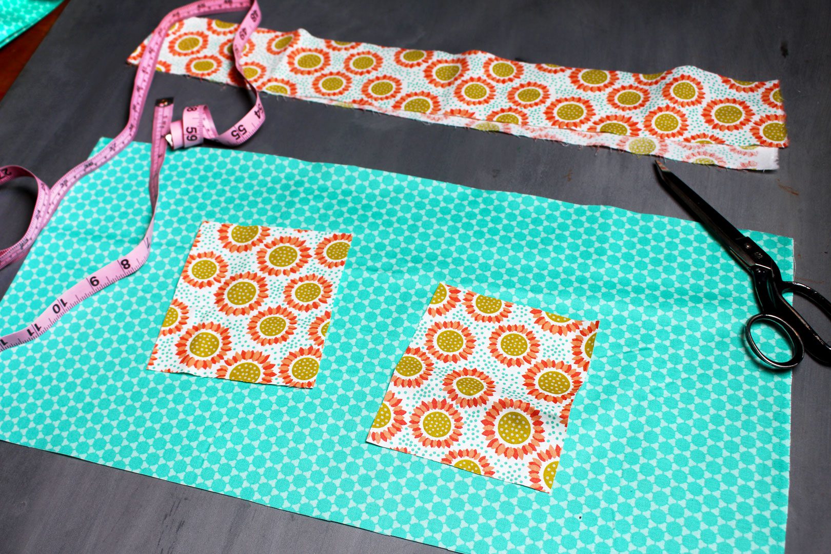Orange floral and teal fabric layed out with a measuring tape and pair of fabric scissors before sewing into an apron pattern. 