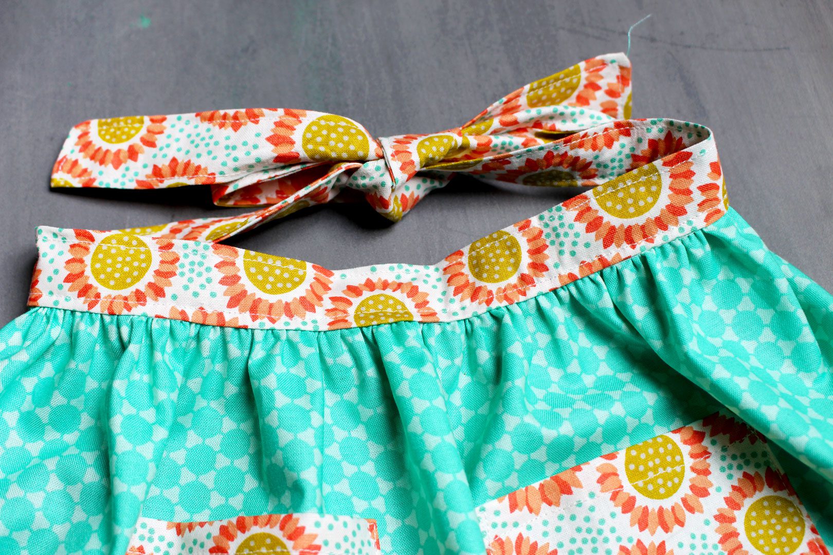 An orange floral waist band for a DIY apron sewed to the top of teal fabric and tied at the back.