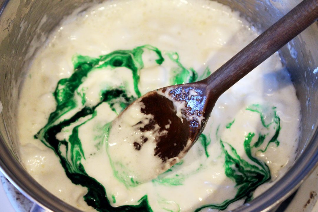 A wooden spoon stirring green food coloring into a mixture of butter and melted marshmallows.