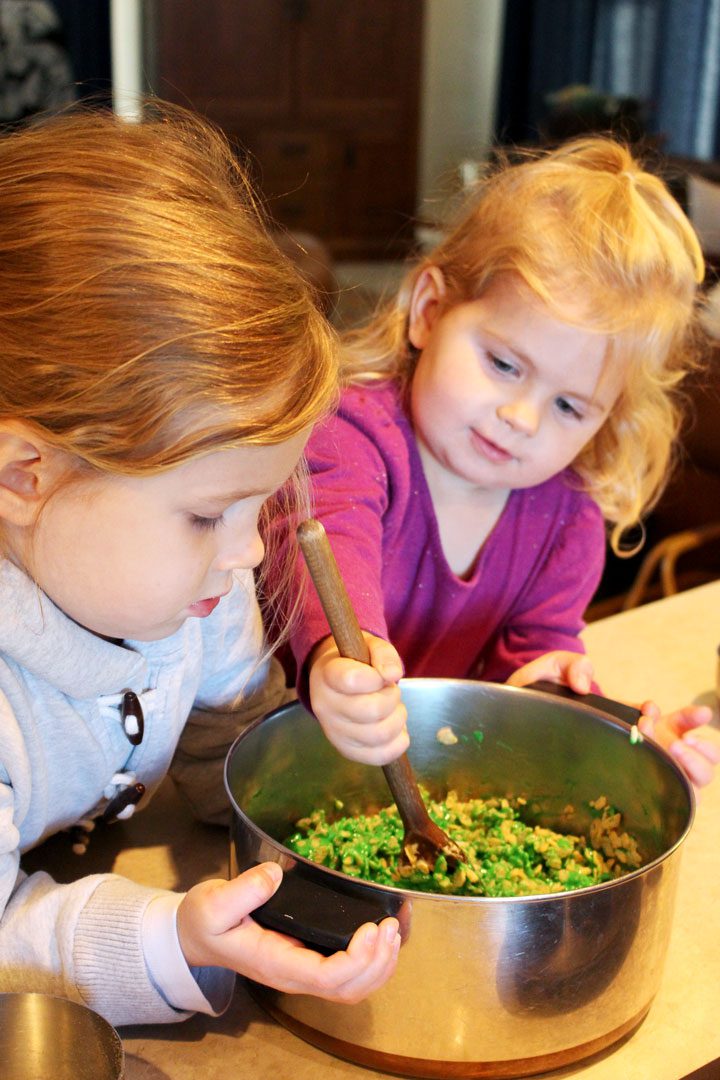 Two girls stirring a green colored crispy rice mix in a large bowl.