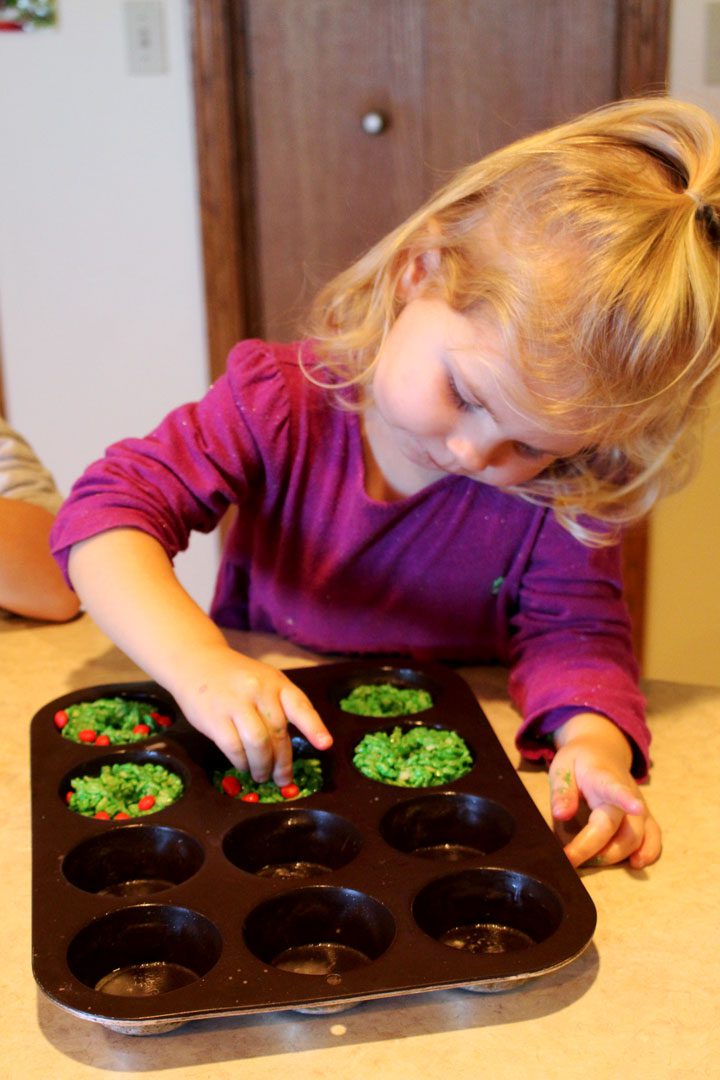 A girl pressing red hot candies into green wreath cookies in a cupcake pan.