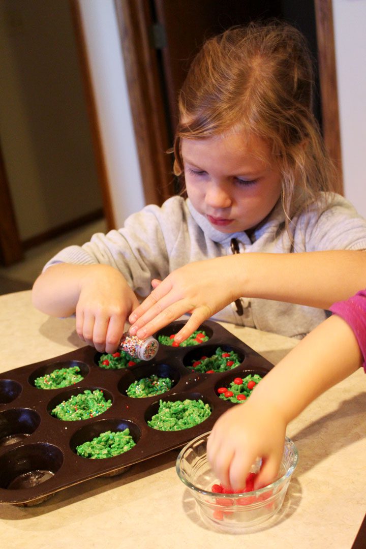 A girl pressing red hot candies into green wreath cookies in a cupcake pan.