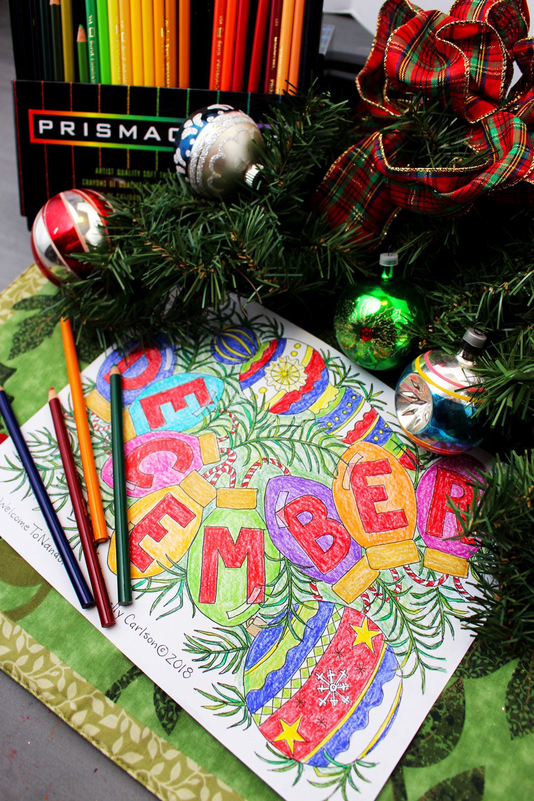 Vertical image of the December coloring page with greenery and colored pencils all around. Nanas Free Tis the Season December Coloring Page is a free downloadable.