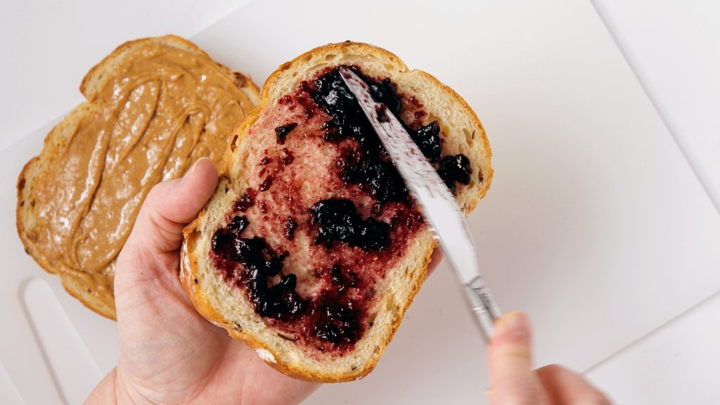 Spreading jelly on bread, with a piece of bread with peanut butter spread in the background.