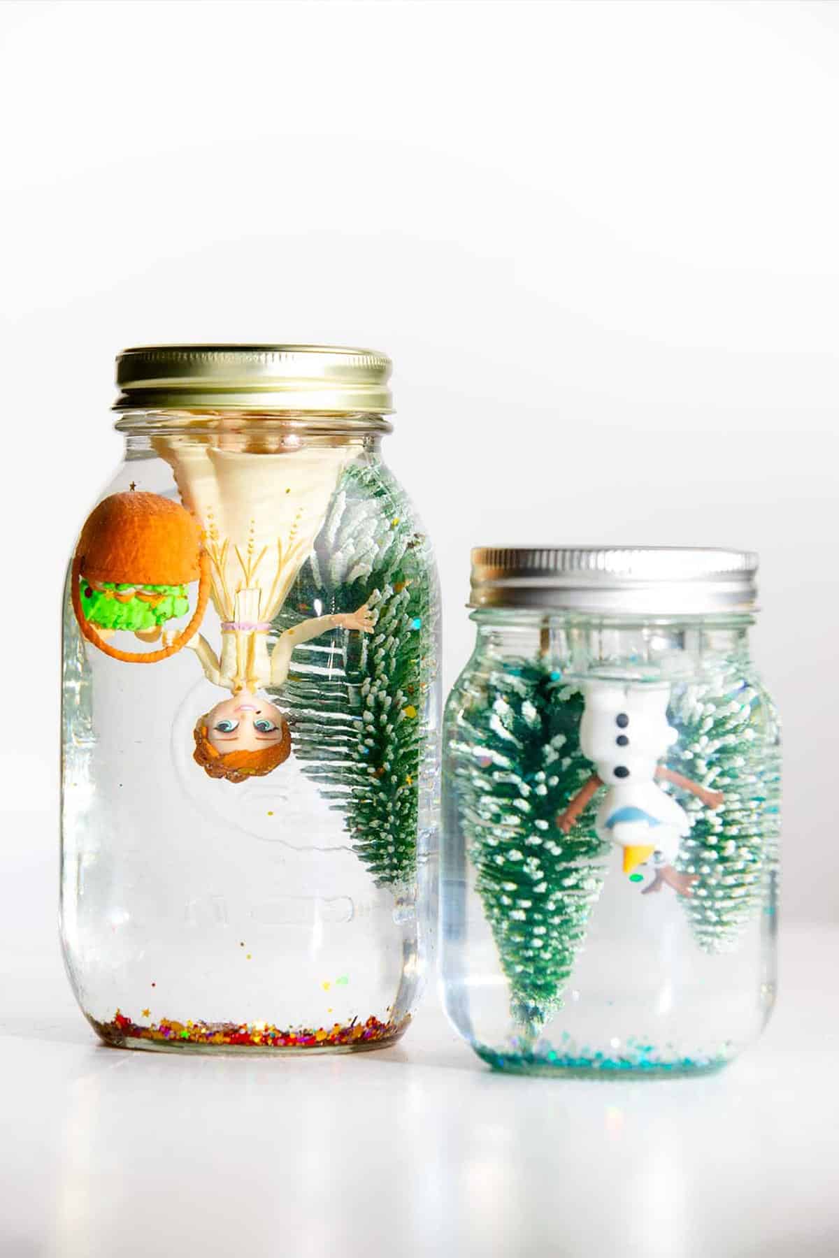 Two mason jar snow globes with trees and Olaf and Anna Frozen characters.