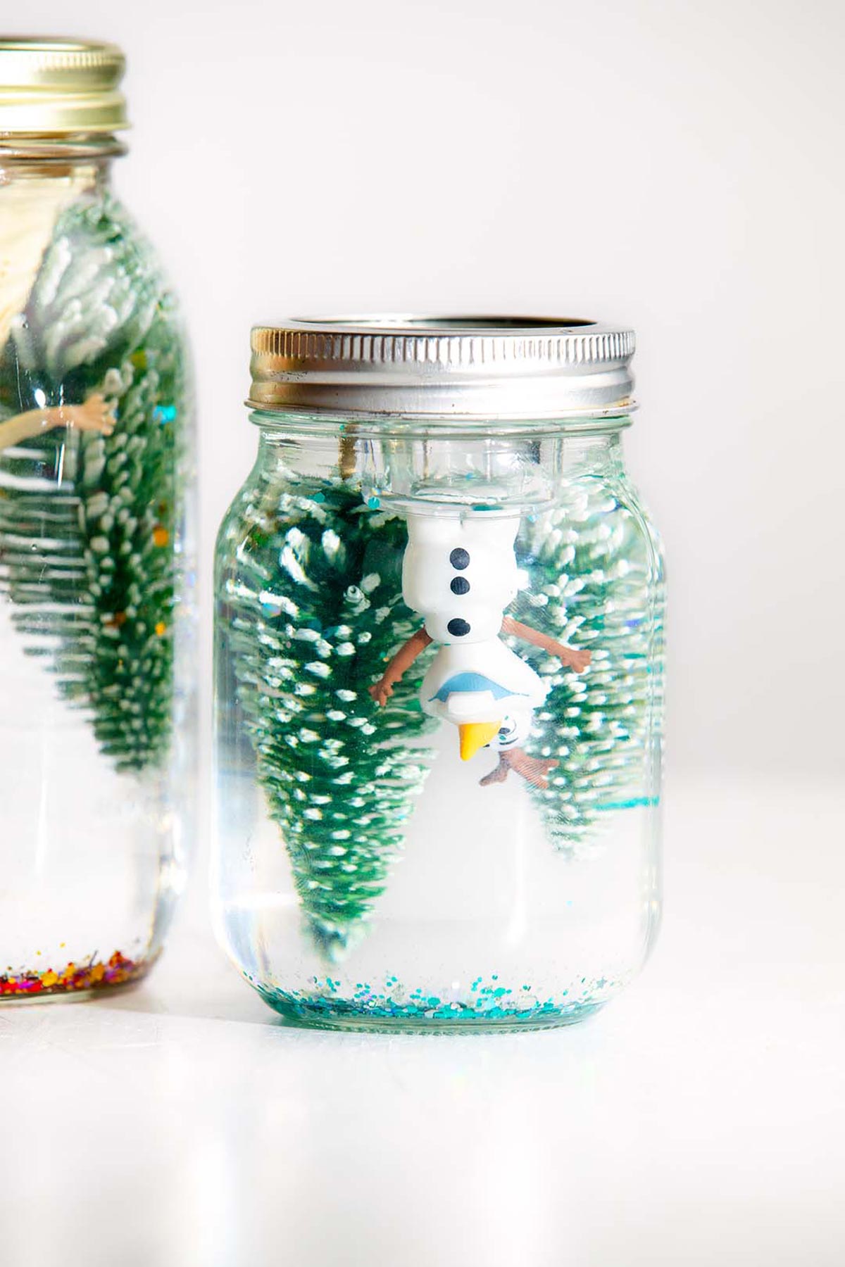 Two mason jar snow globes with trees and Olaf and Anna Frozen characters.