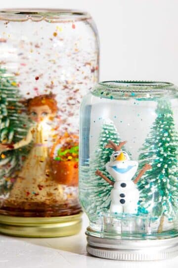 Two mason jar snow globes with trees and Olaf and Anna Frozen characters.