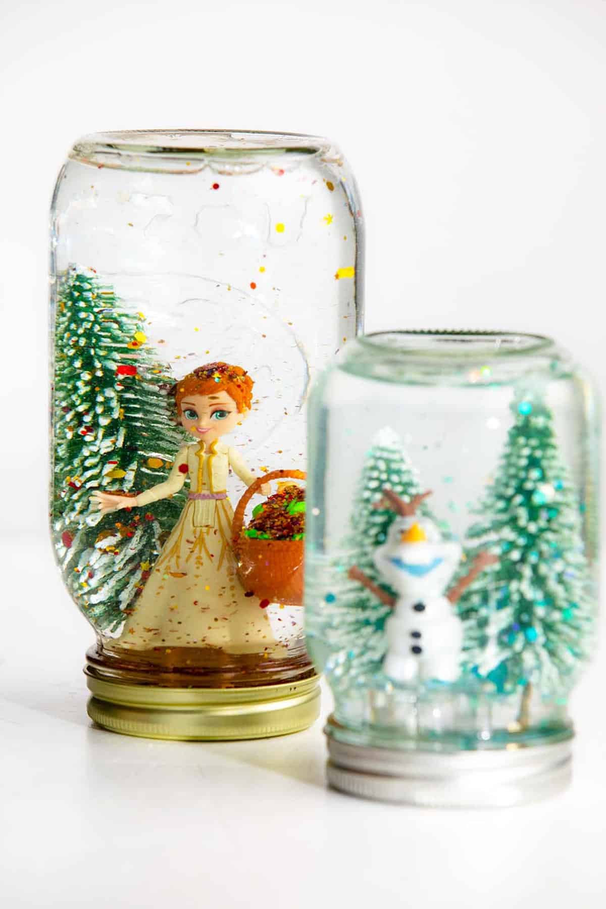 Two mason jar snow globes with trees and Olaf and Anna Frozen characters.