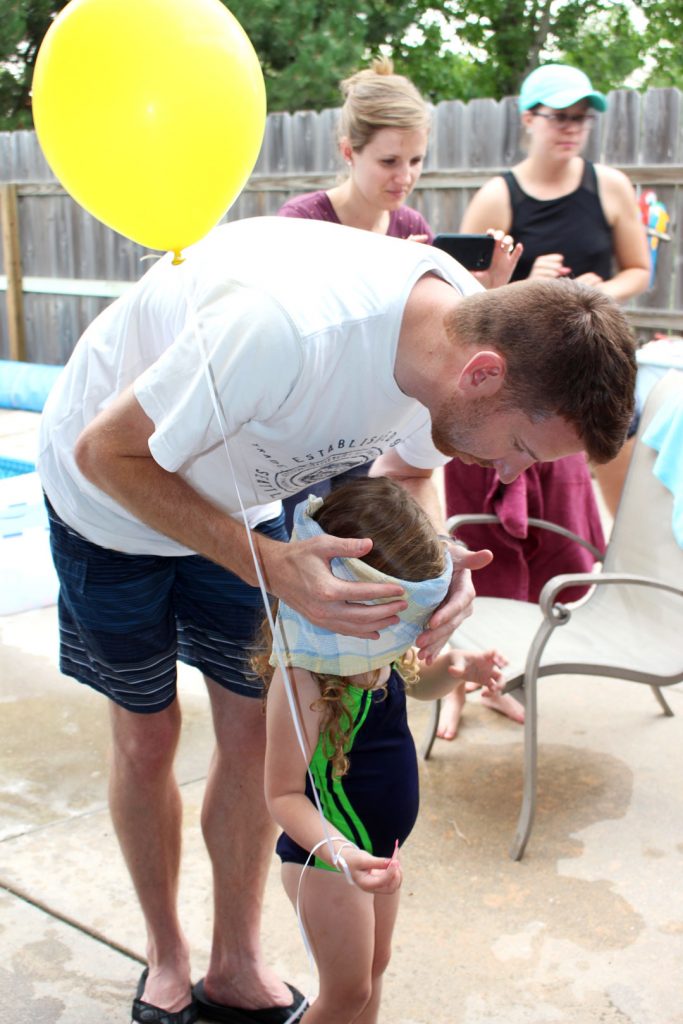 A child with a balloon being blindfolded by a parent to play a pin the tail game at a party.