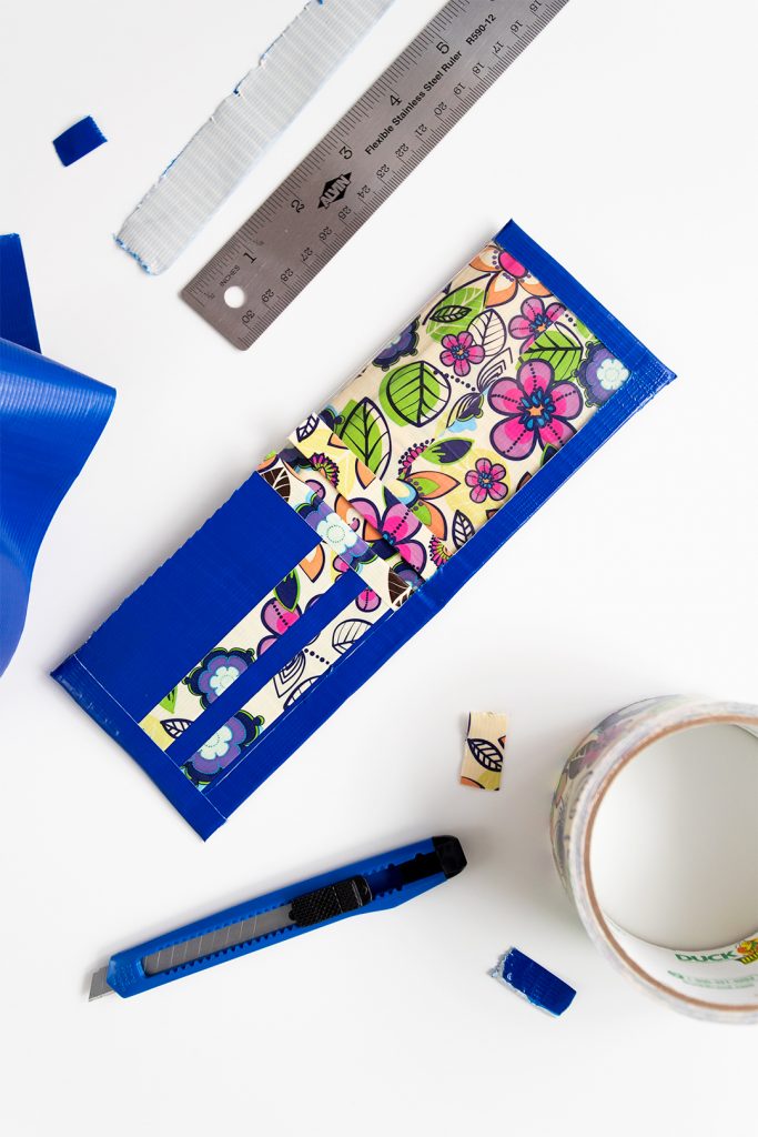A flower patterned duct tape wallet, utility knife and blue roll of duct tape, ruler, pieces of duct tape.