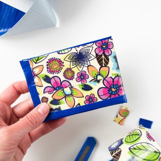 A hand holding a flower patterned duct tape wallet, utility knife and roll of blue duct tape in the background.