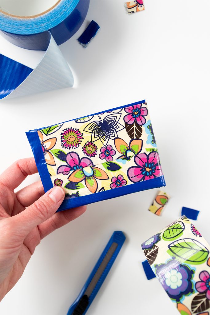 A hand holding a flower patterned duct tape wallet, utility knife and roll of blue duct tape in the background.