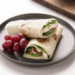 A turkey wrap with greens and bacon on a plate with grapes.