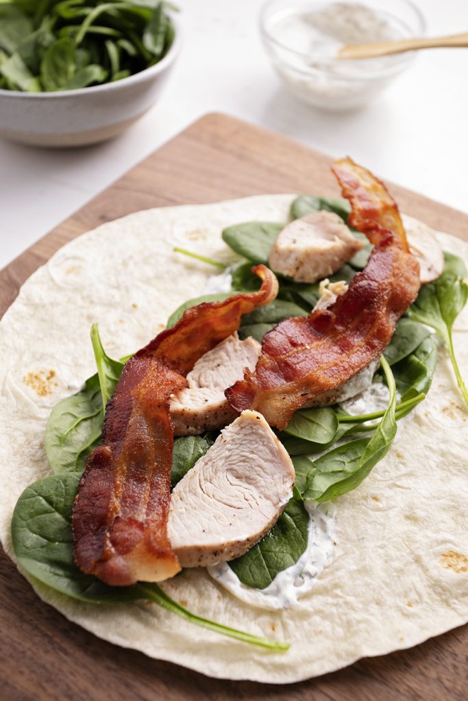 A tortilla, dressing, spinach, bacon, and turkey on a cutting board.