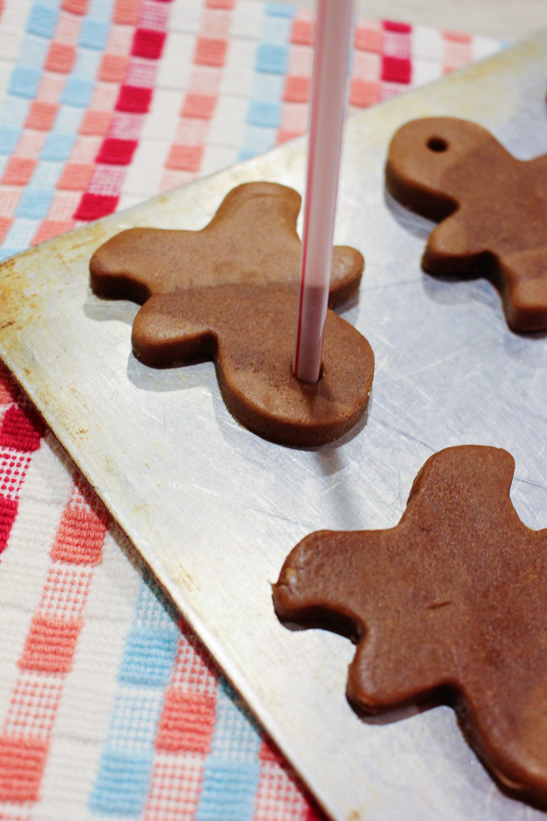 A straw poking a hole in the top of a gingerbread cookie sitting on a baking sheet.