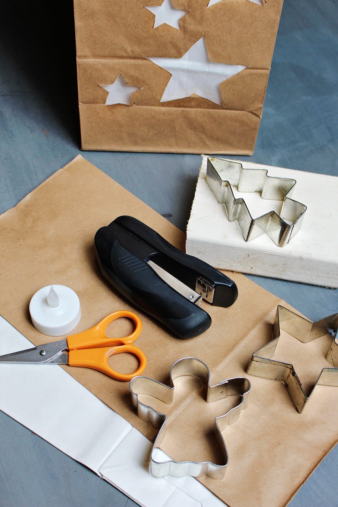 A pair of scissors, stapler, tea light, and cookie cutters sitting near a brown paper bag Christmas luminary.