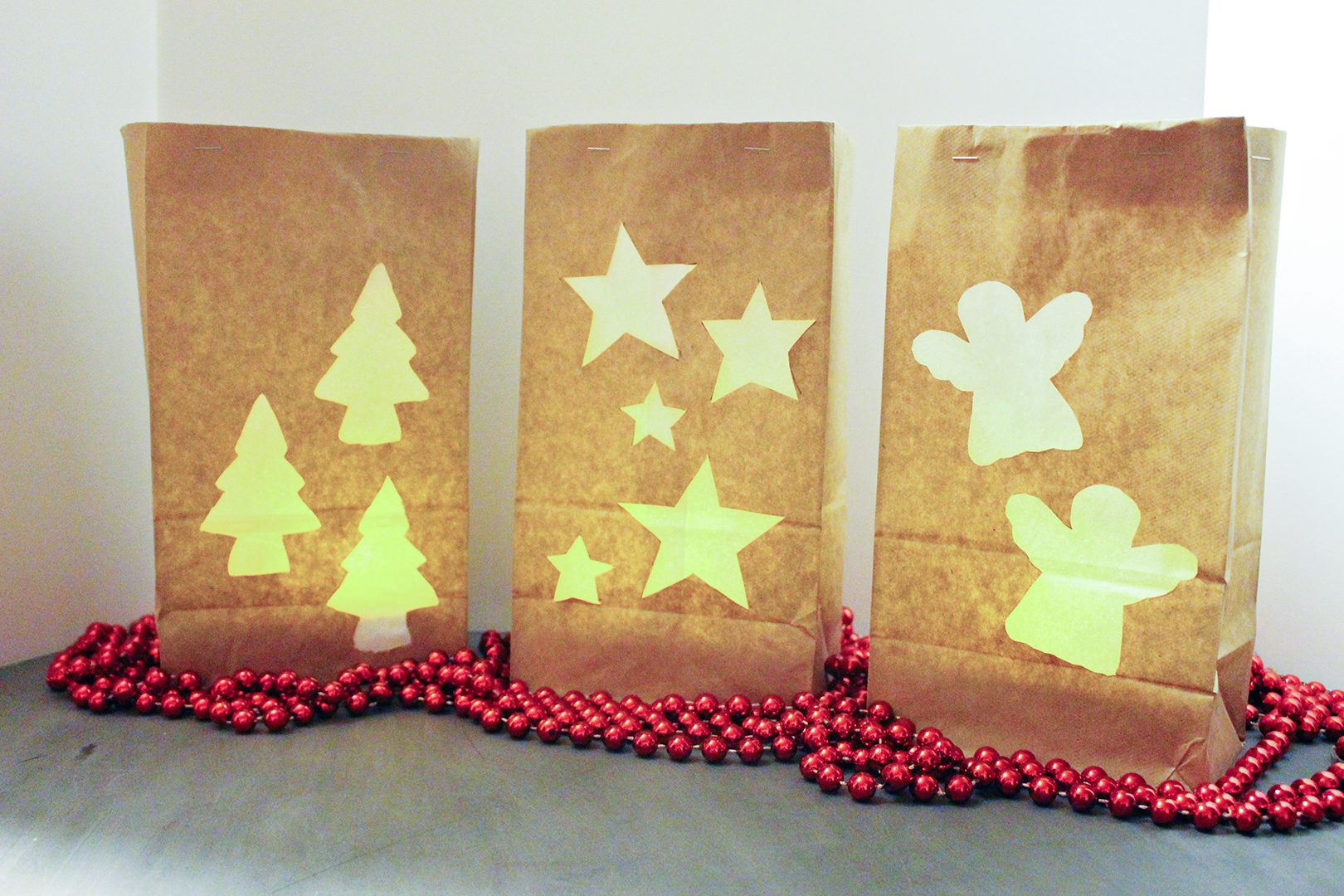 Three brown paper bag luminaries with angel, star, and tree cutouts, surrounded by red Christmas beads.