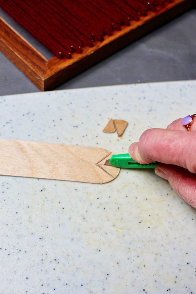 A utility knife cutting a craft stick into a heddle.