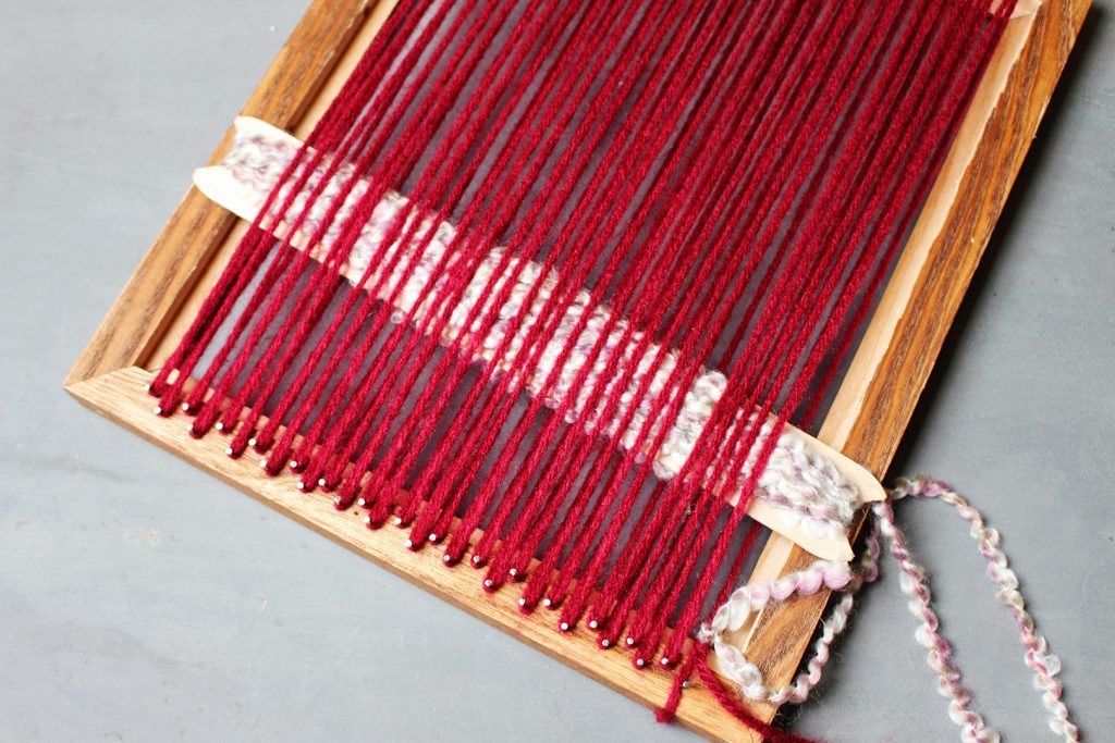 A heddle weaving through the warp yarns on a wooden picture frame loom.