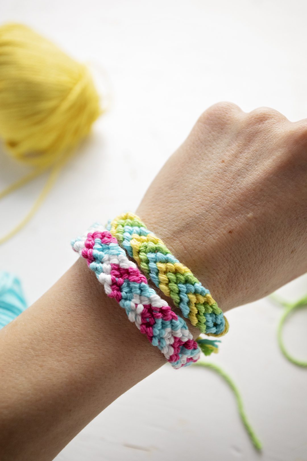 Two chevron pattern bracelets in green, blue, yellow, pink, and white embroidery floss, tied on a person's wrist.