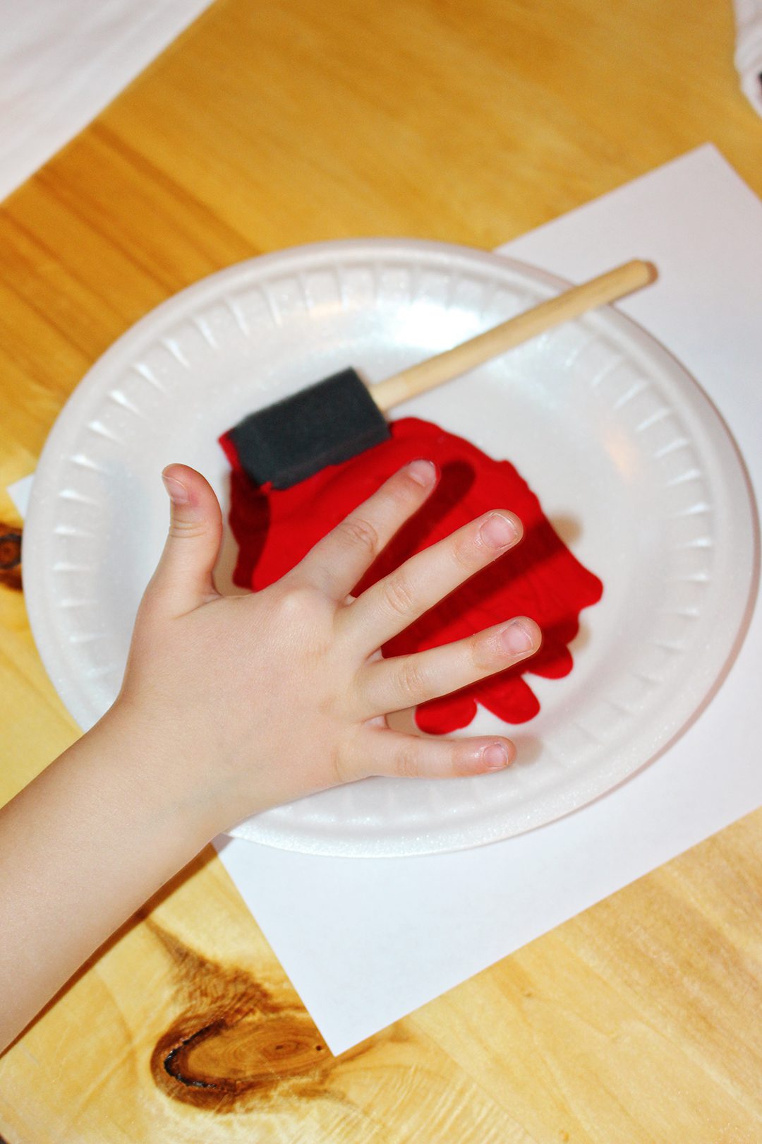 A child touching red paint spread on a plastic plate with a sponge brush.