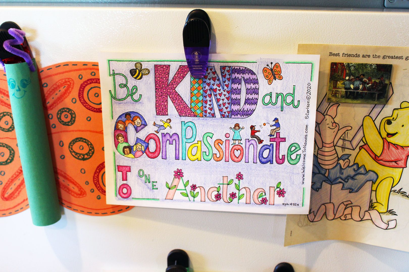 A coloring page with the words "Be Kind and Compassionate To one Another", colored by colored pencils, hanging on a refrigerator.