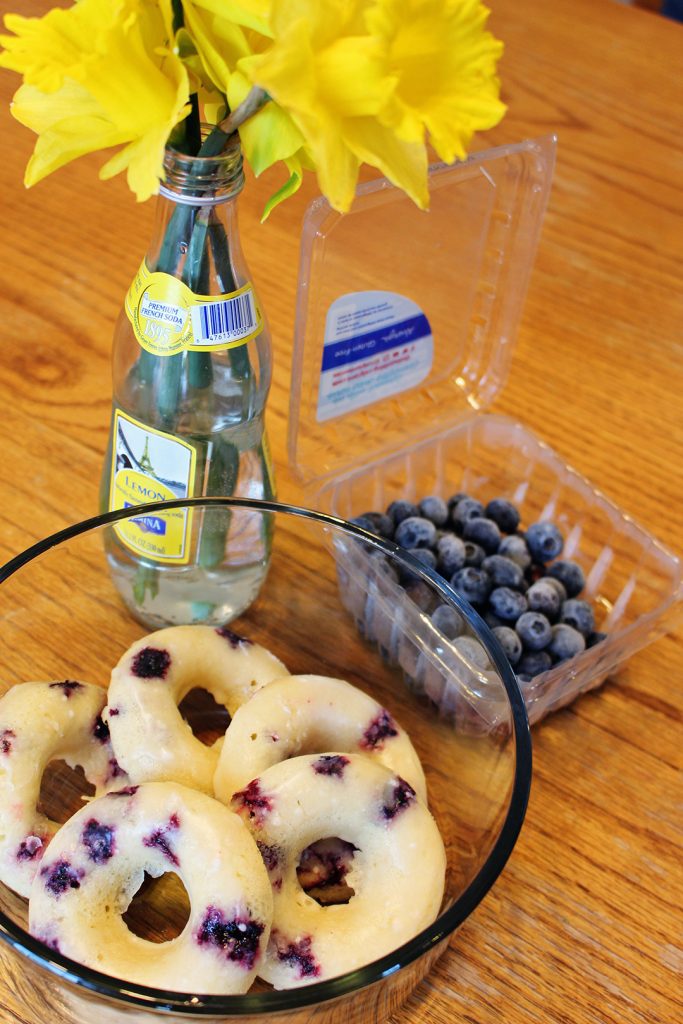 A bowl of Lemon Blueberry Donuts with flowers and blueberries nearby.