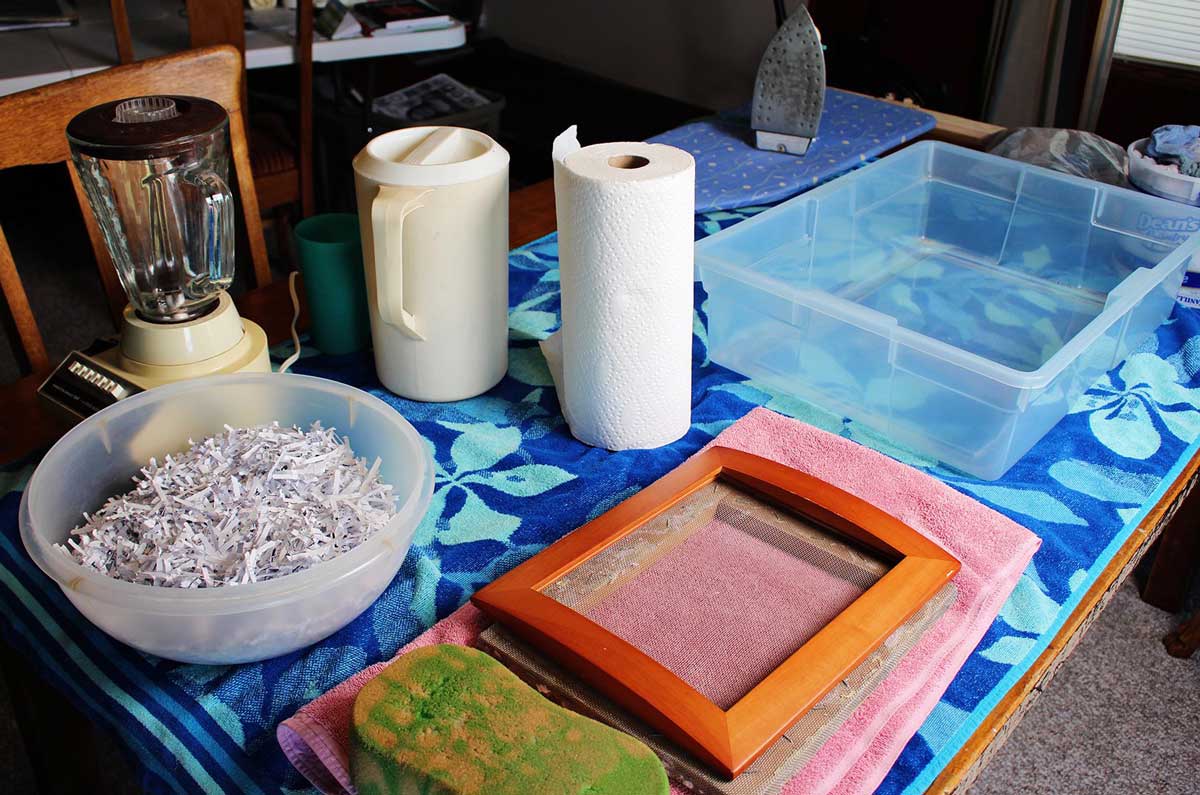 A bowl of paper scraps, a picture frame mould and deckle, paper towels, a beach towel, a tub of water, a picture, and an iron.