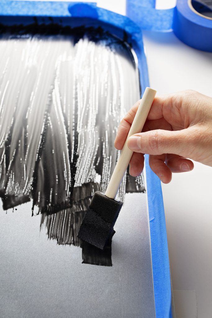 A sponge brush painting chalkboard paint onto a metal tray with paint tape edging.