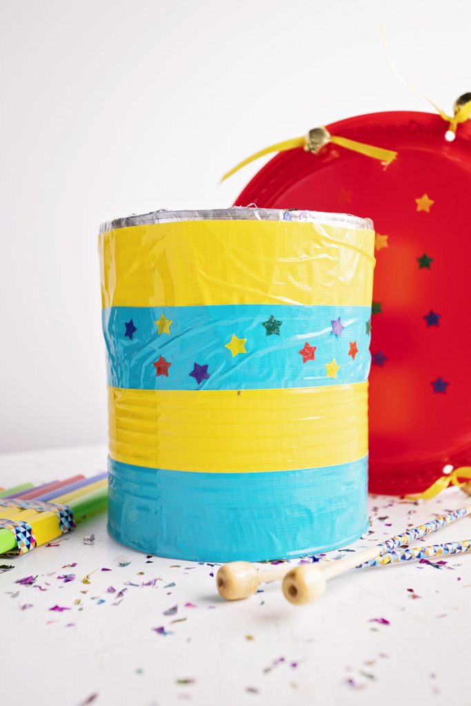 A striped can made into a drum, decorated with stickers, next to a tambourine, drumsticks, and a fife.