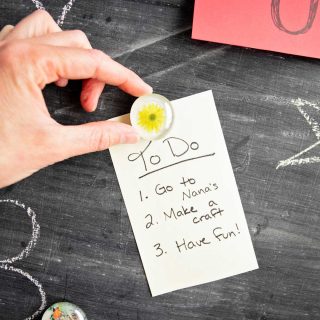A hand placing a glass gem flower magnet on a chalkboard to hold up a To Do list.