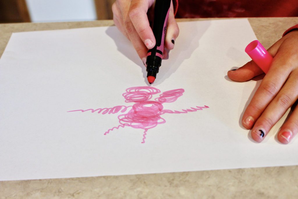 A child coloring a pink scribble monster with a marker on a piece of paper