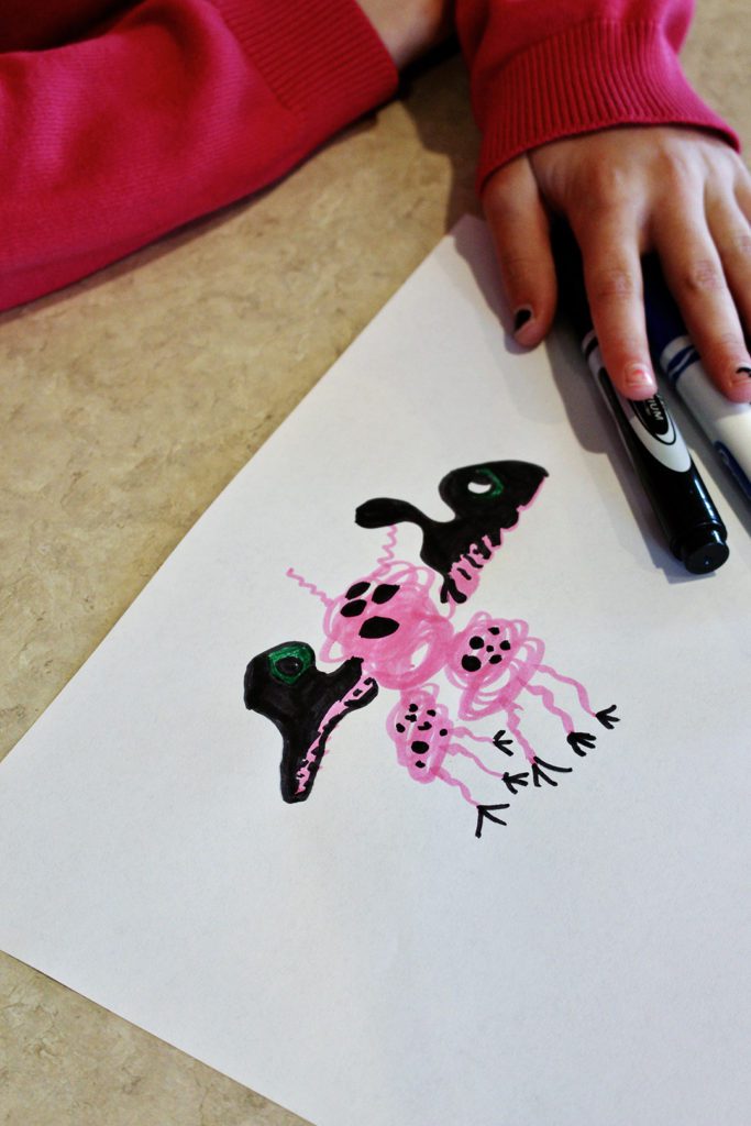 A child coloring a pink scribble monster with a marker on a piece of paper